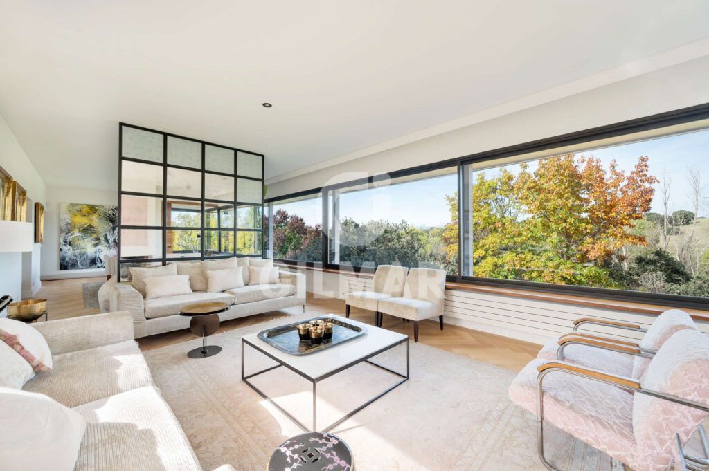 Salón moderno con sofás en tonos neutros y ventanales panorámicos en chalet en Madrid en Ciudalcampo.