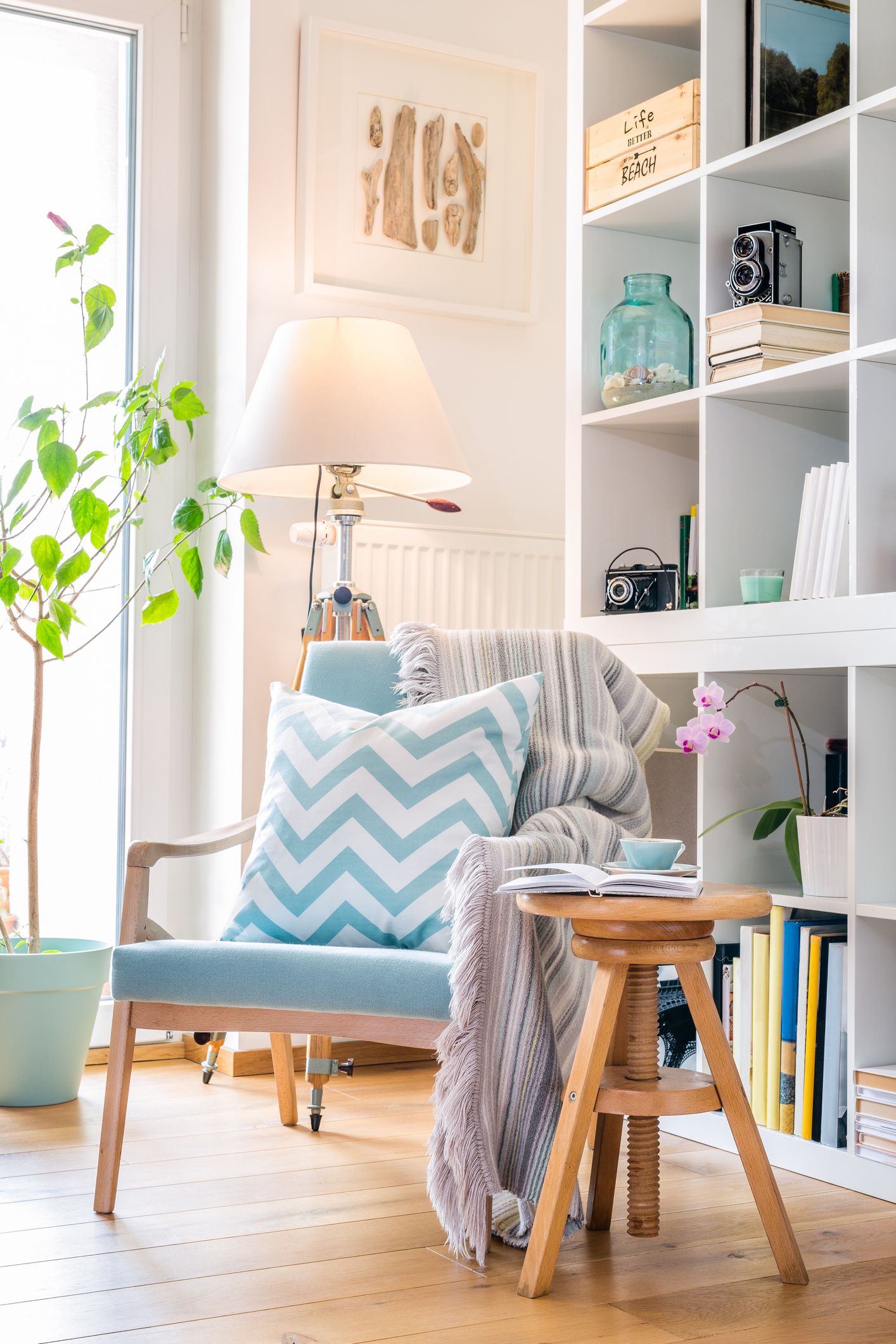 Rincón de lectura en casa con butaca azul, cojín decorativo, manta, lámpara de pie y estantería junto a una ventana con luz natural